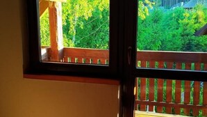Una habitación con piso de madera, un ventanal con vista a un balcón y follaje, y una puerta con manija.