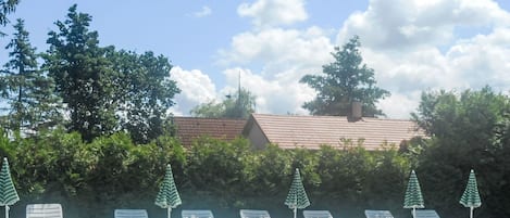 A swimming pool with white lounge chairs and green umbrellas, surrounded by trees and a building with a tiled roof.
