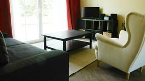 Una sala de estar con un sofá, una butaca, una mesa de centro, un mueble para televisión con televisor y parlantes, y una ventana con cortinas.