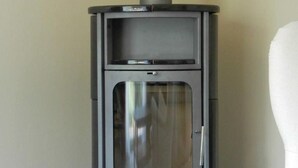Una estufa de leña moderna y compacta con puerta de cristal, ubicada frente a una pared lisa.