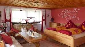 Ein gemütliches Zimmer mit einem hölzernen Bett, einer Holz-Couch und einem Holztisch. Es gibt ein Fenster mit Vorhängen, eine Wand mit einem dekorativen Schild und einen kleinen Tisch mit Blumen.