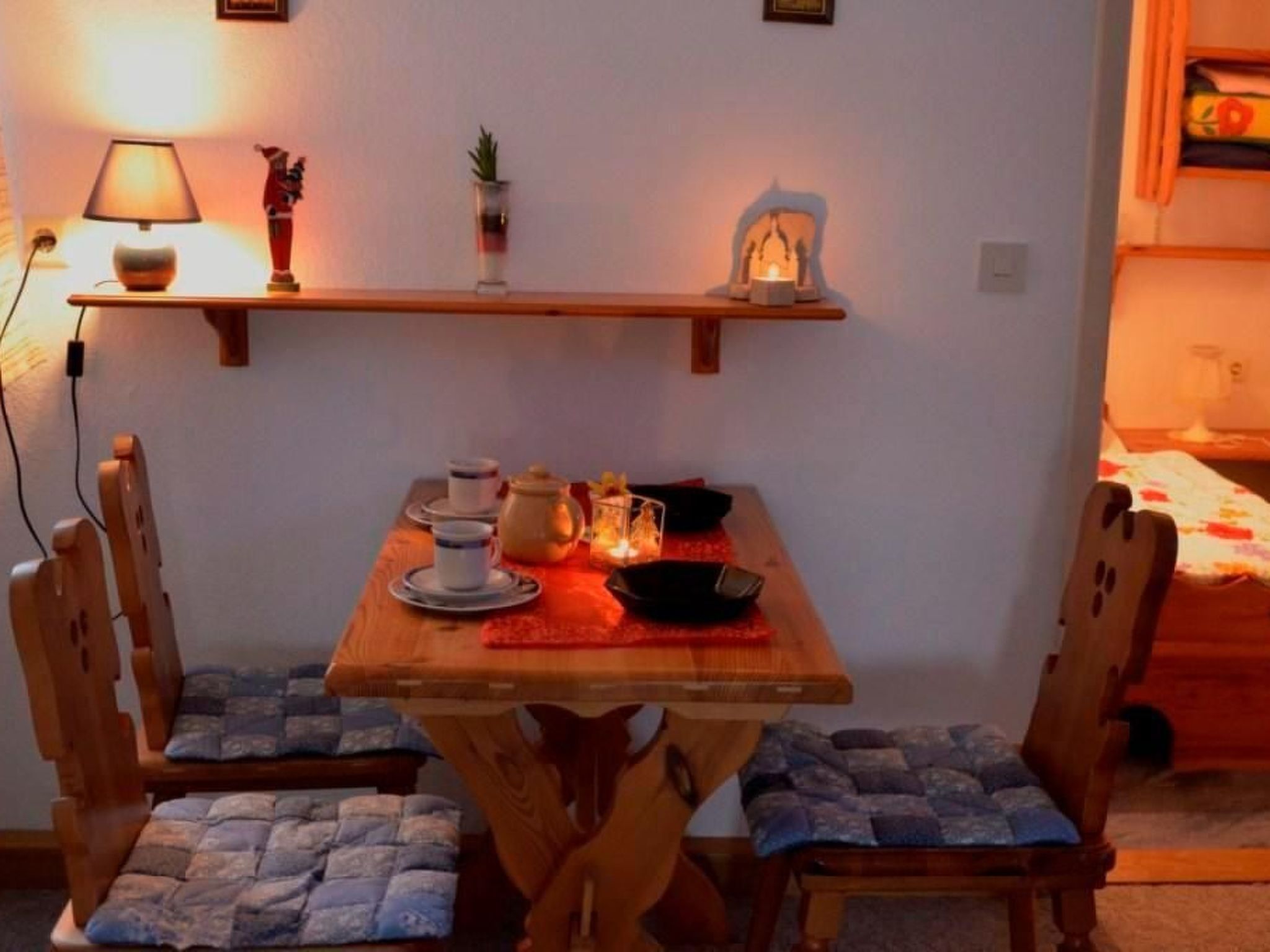 A cozy dining area with a wooden table, two chairs, a lamp, a small shelf with decorative items, and a lit candle.
