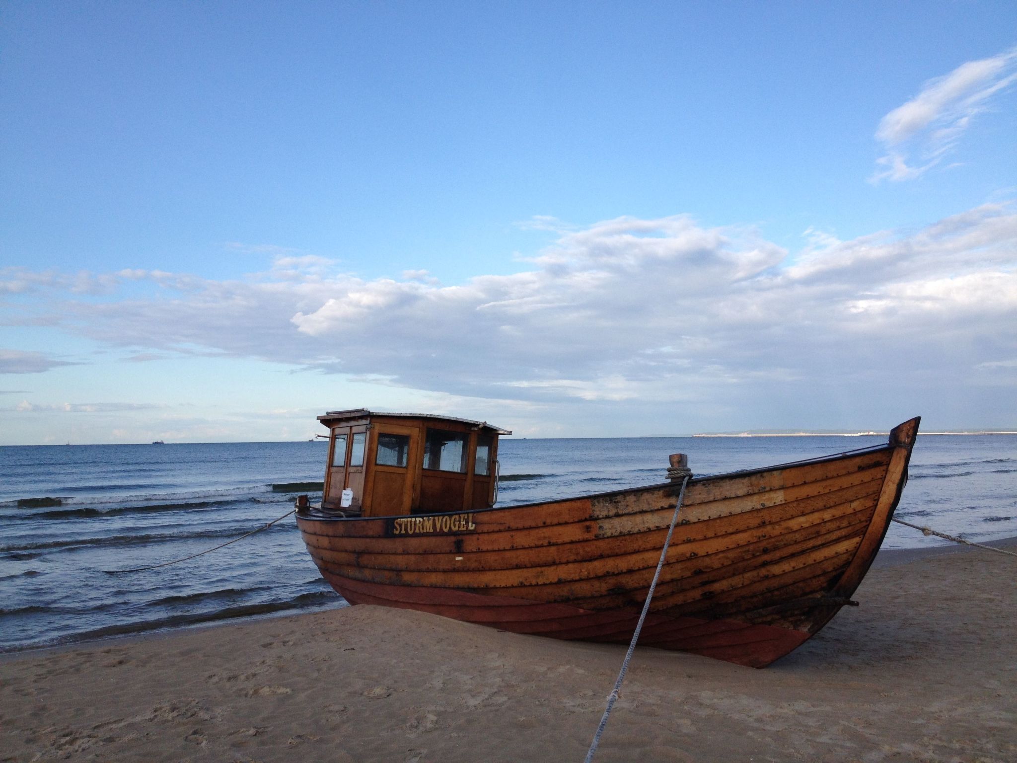 A wooden boat named STURMVOGEL on a sandy beach with calm water and a clear sky.