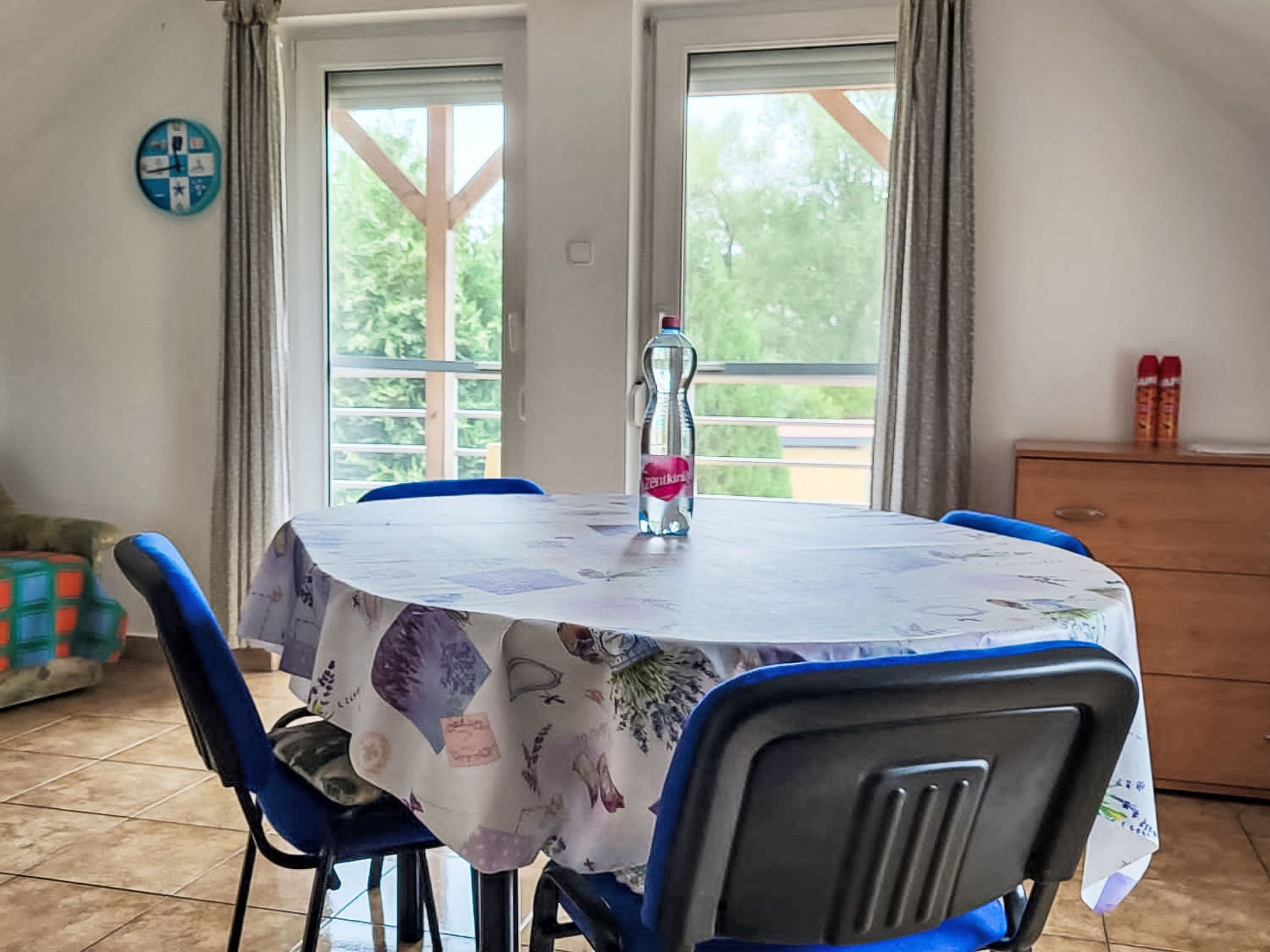 A dining area with a round table covered by a tablecloth with a floral pattern, surrounded by blue chairs. A bottle of water is placed on the table.