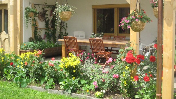Un espacio al aire libre cubierto con una mesa y sillas de madera, rodeado de diversas plantas en flor y cestas colgantes.