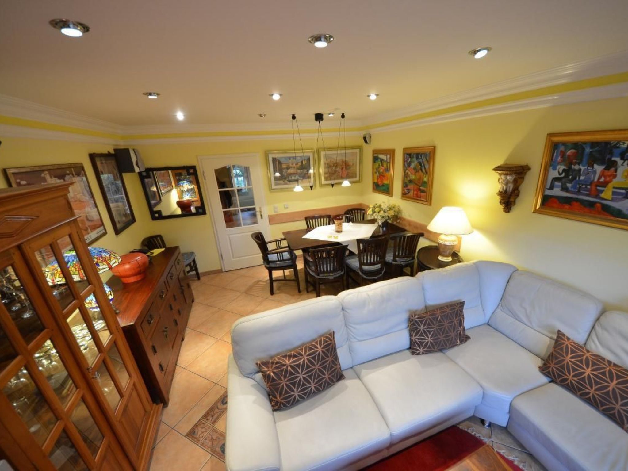 A living room with a white sofa, dining table, and chairs. There is a wooden cabinet with glass doors and a lamp on the dining table.