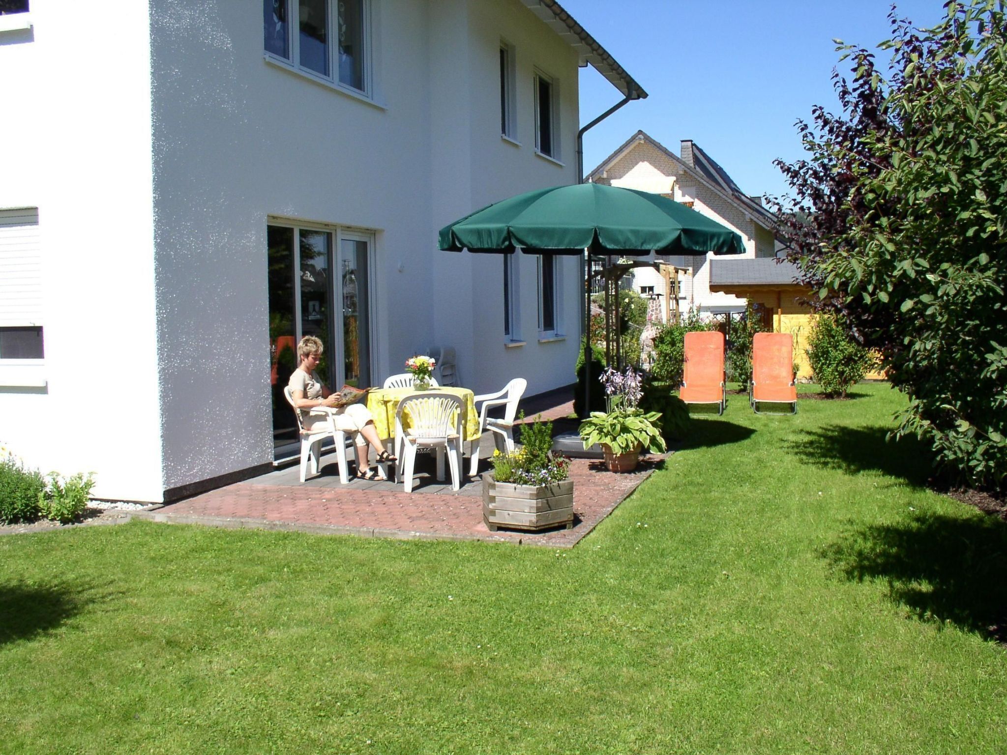 A patio with a table and chairs, a green umbrella, and a garden with a lawn and plants.