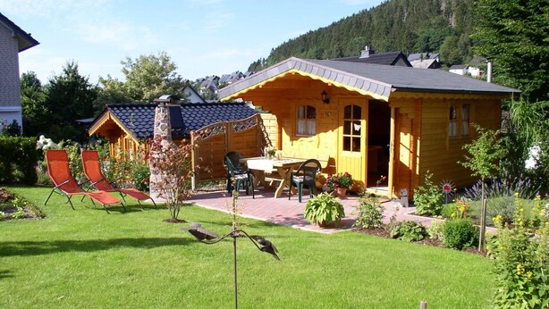 A yellow wooden house with a stone chimney, a patio with a table and chairs, and a well-maintained garden with grass and plants. - Apartment Wiese by Interhome (Bruchhausen)