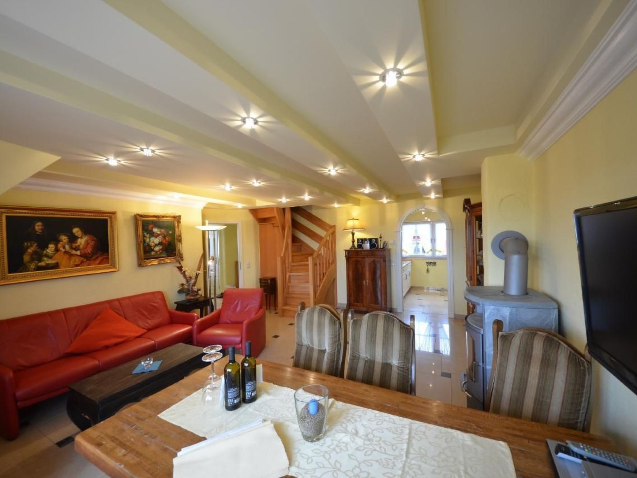 A living room with a red sofa, a wooden coffee table with wine bottles, and a TV on the right. There is a staircase leading to another room.