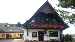 Ein zweigeschossiges Gebäude mit einem geneigten Dach, einem Balkon mit roten Blumen und einer kleinen Veranda mit Sitzgelegenheiten.
