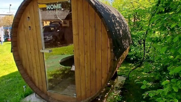 Een houten, vatvormige constructie met de tekst 'HOME DELUXE', een zichtbaar interieur en een kleine tuin eromheen.