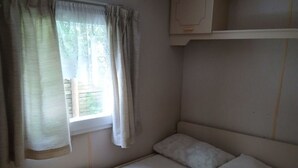 Een kleine kamer met een enkel bed, een raam, gordijnen, een plafondventilator en een plank boven het bed.