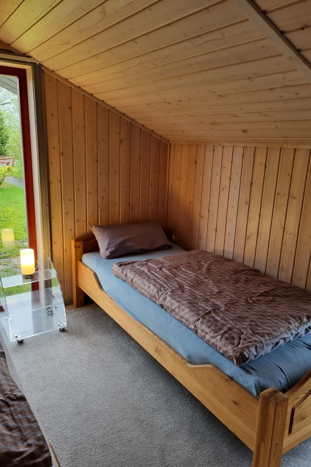 A single wooden bed with a blue mattress and a patterned blanket, a clear glass table with a candle, and a view of greenery outside.
