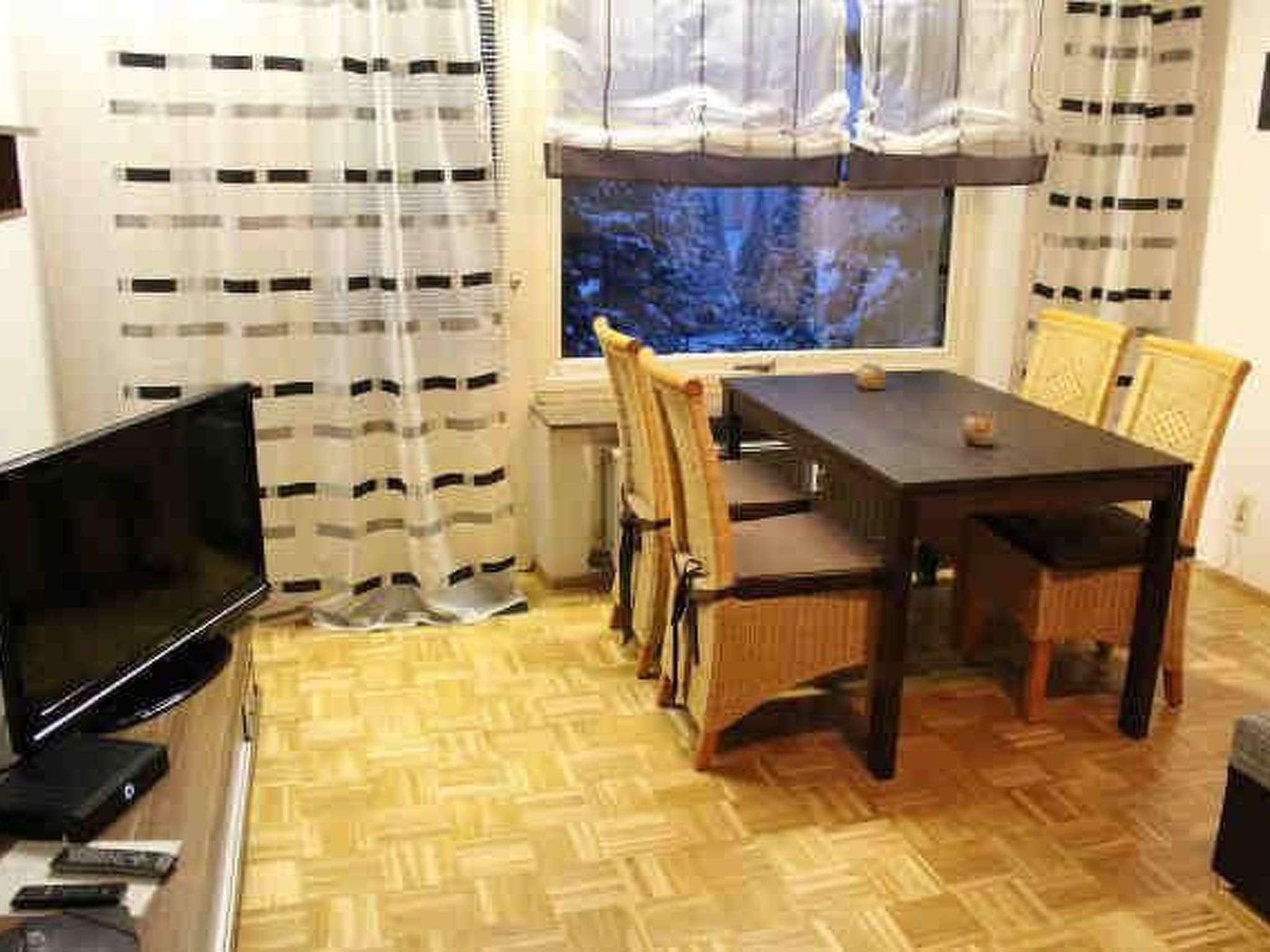 A small room with a wooden floor, a TV on a stand, a dining table with chairs, and a window with curtains.