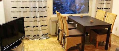 A small room with a wooden floor, a TV on a stand, a dining table with chairs, and a window with curtains.