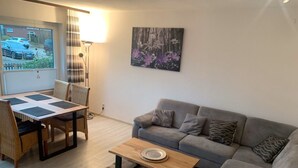 A living room with a grey sofa, a wooden coffee table, a dining table with chairs, and a painting on the wall.