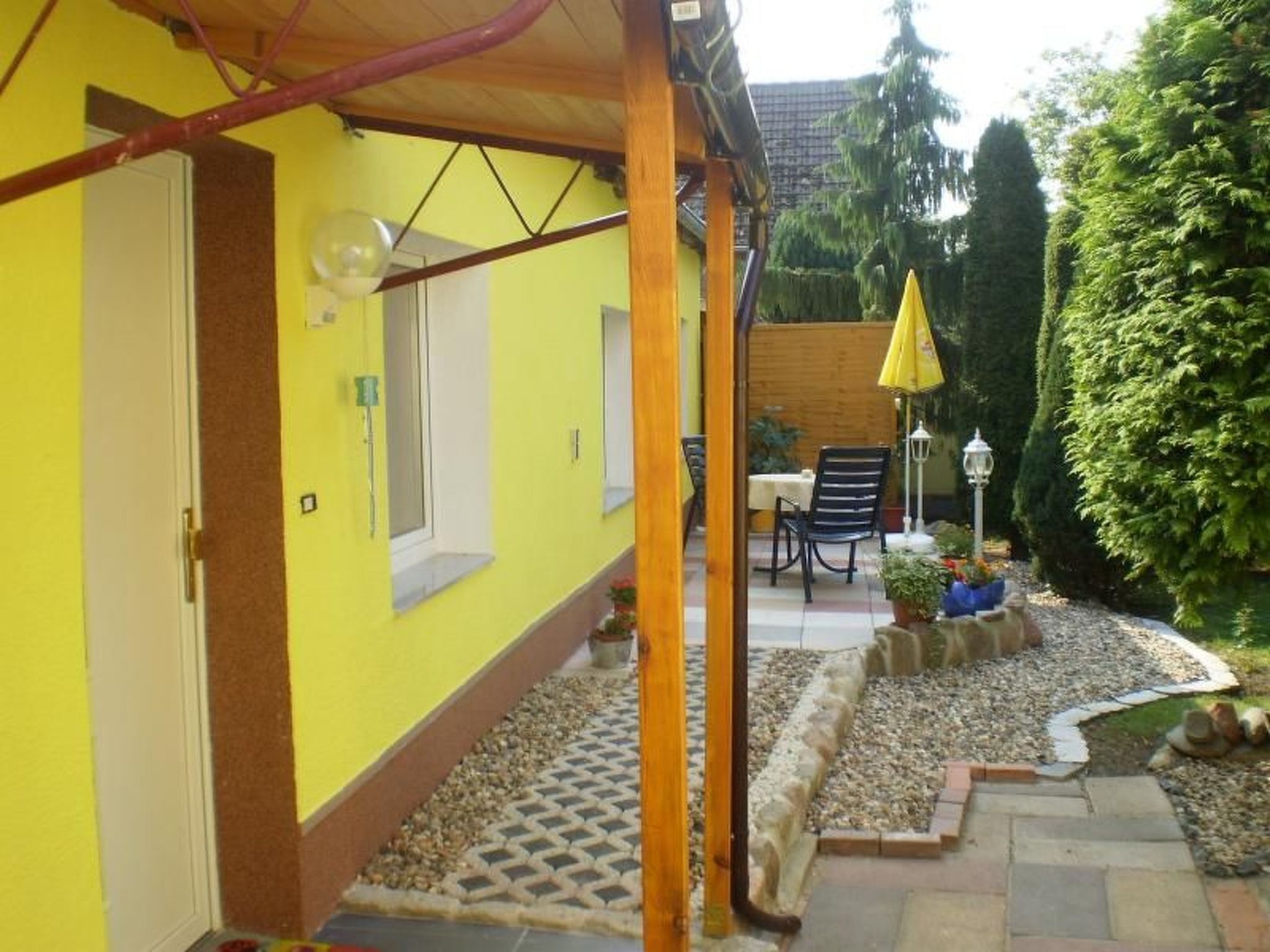 A yellow building with a covered patio area, a table with chairs, and a garden with a lamp post.