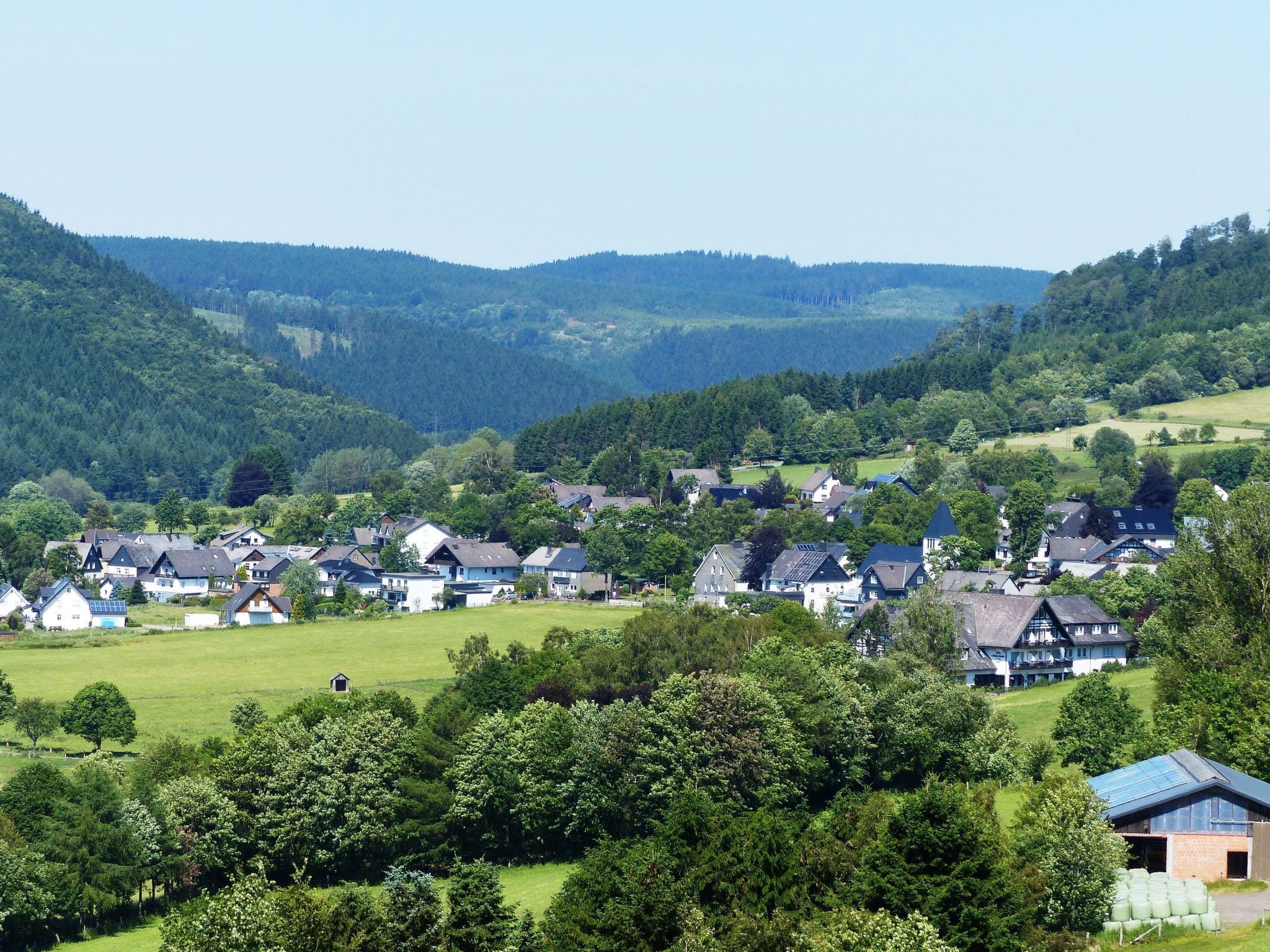 A rural village with houses surrounded by lush greenery and hills in the background.