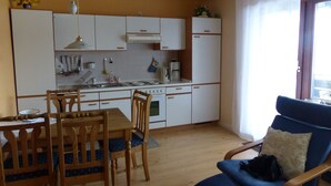 Kompakti keittiö, jossa on valkoiset kaapit, ruokapöytä tuoleineen sekä sininen nojatuoli, jossa on tyyny.