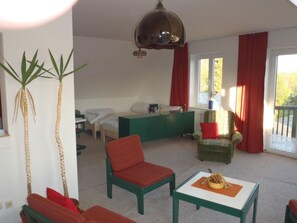 Et værelse med en seng, en grøn lænestol, en rød lænestol, et lille bord med snacks og et stort vindue med røde gardiner.