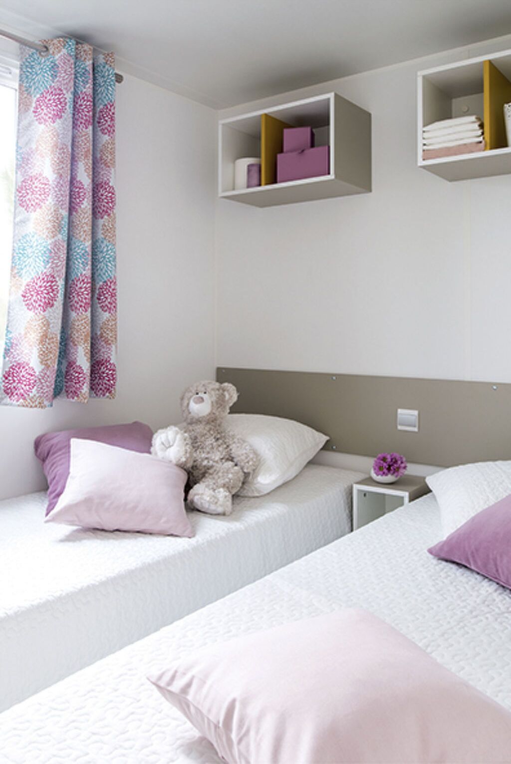 A bedroom with two beds, a teddy bear, and a shelf with books and a vase.