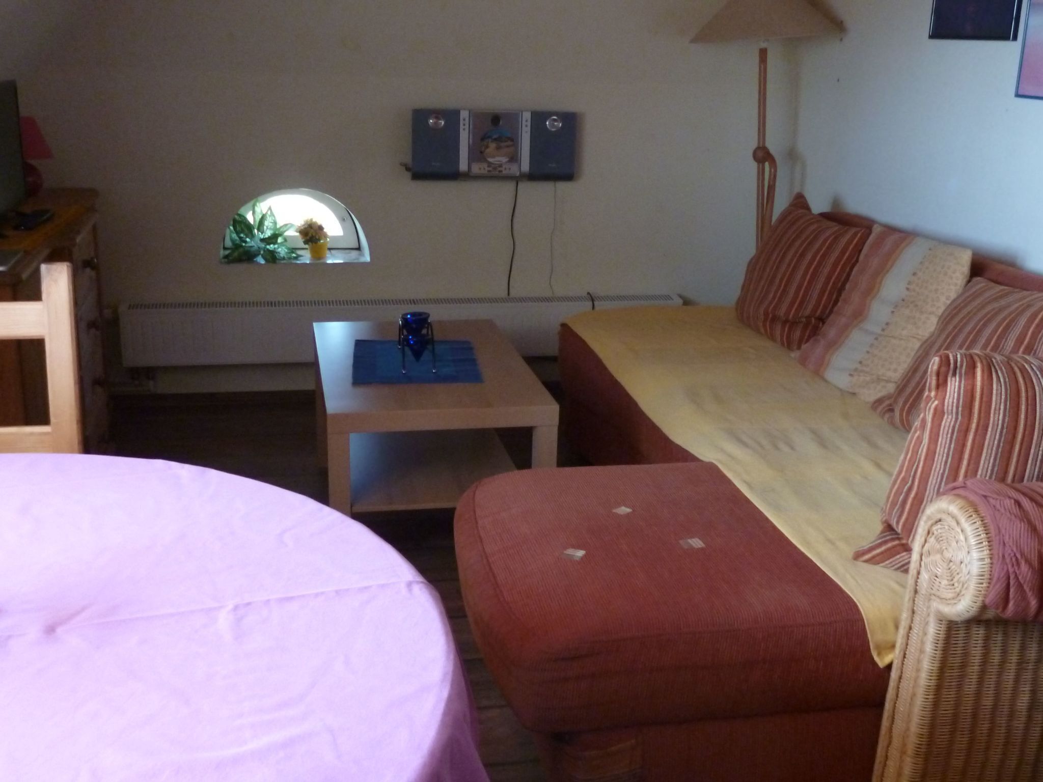 A small, cozy room with a single bed, a sofa, and a table. There is a window with a view of the outdoors, and a wall-mounted heater.