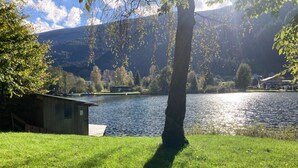 Blick auf einen See mit einer Holzsteganlage und einem kleinen Gebäude, umgeben von üppiger Vegetation und Bergen im Hintergrund.