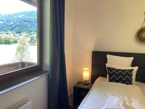 Ein Hotelzimmer mit Bett, Nachttisch mit Lampe, Fenster mit Vorhängen und Blick auf See und Wald.