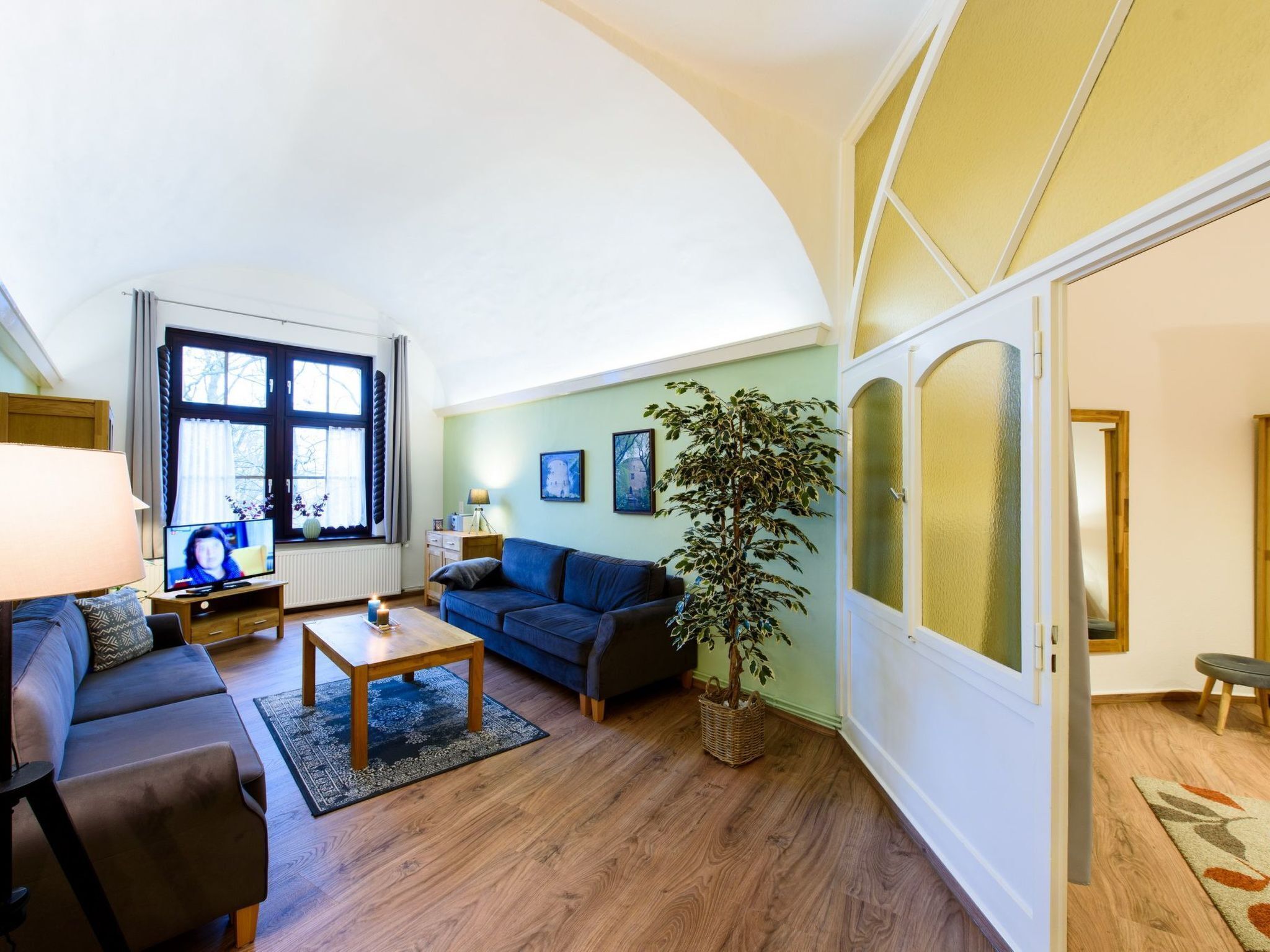 Ein Wohnzimmer mit einer Couch, einem Couchtisch und einem Fernseher. Ein Fenster mit Vorhängen und einer Pflanze ist ebenfalls vorhanden.