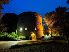 Une tour de pierre historique, dotée d’une tourelle ronde, de fenêtres illuminées et d’un espace d’entrée pavé, le tout entouré d’arbres.