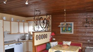 Een kleine keuken met een spoelbak, kookplaat en kastjes. Er is een eethoek met een tafel, stoelen en een bankje.