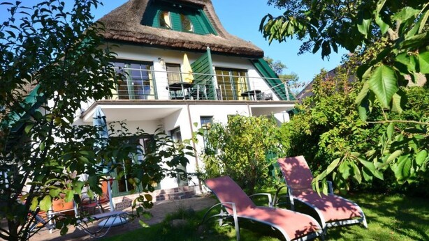 Ein zweigeschossiges Gebäude mit einem Strohdach, einem Balkon mit Sitzgelegenheiten im Freien und einem gepflegten Garten mit Liegestühlen.