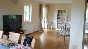 Een ruime woonkamer met een houten plafond, een televisie, een eettafel met stoelen, en een witte kastenwand.