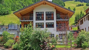 Un chalet de dos pisos con fachada de madera, varios balcones y vista a colinas verdes y montañas al fondo.
