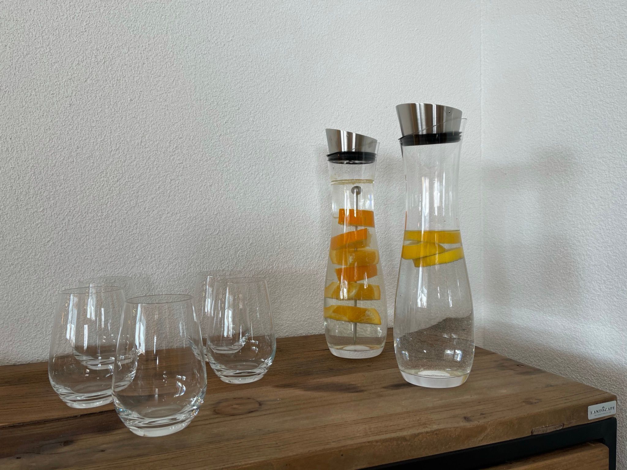 Dos botellas de vidrio con cáscaras de naranja y tapones metálicos, y tres vasos de vidrio vacíos sobre una repisa de madera.