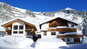 Un chalet de madera de dos pisos con techo nevado, rodeado por un paisaje montañoso cubierto de nieve.