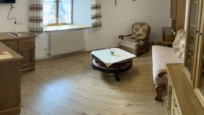 Um quarto aconchegante com piso de madeira, uma mesa redonda com toalha branca, um sofá, um armário de madeira, uma cadeira e uma televisão fixada na parede.