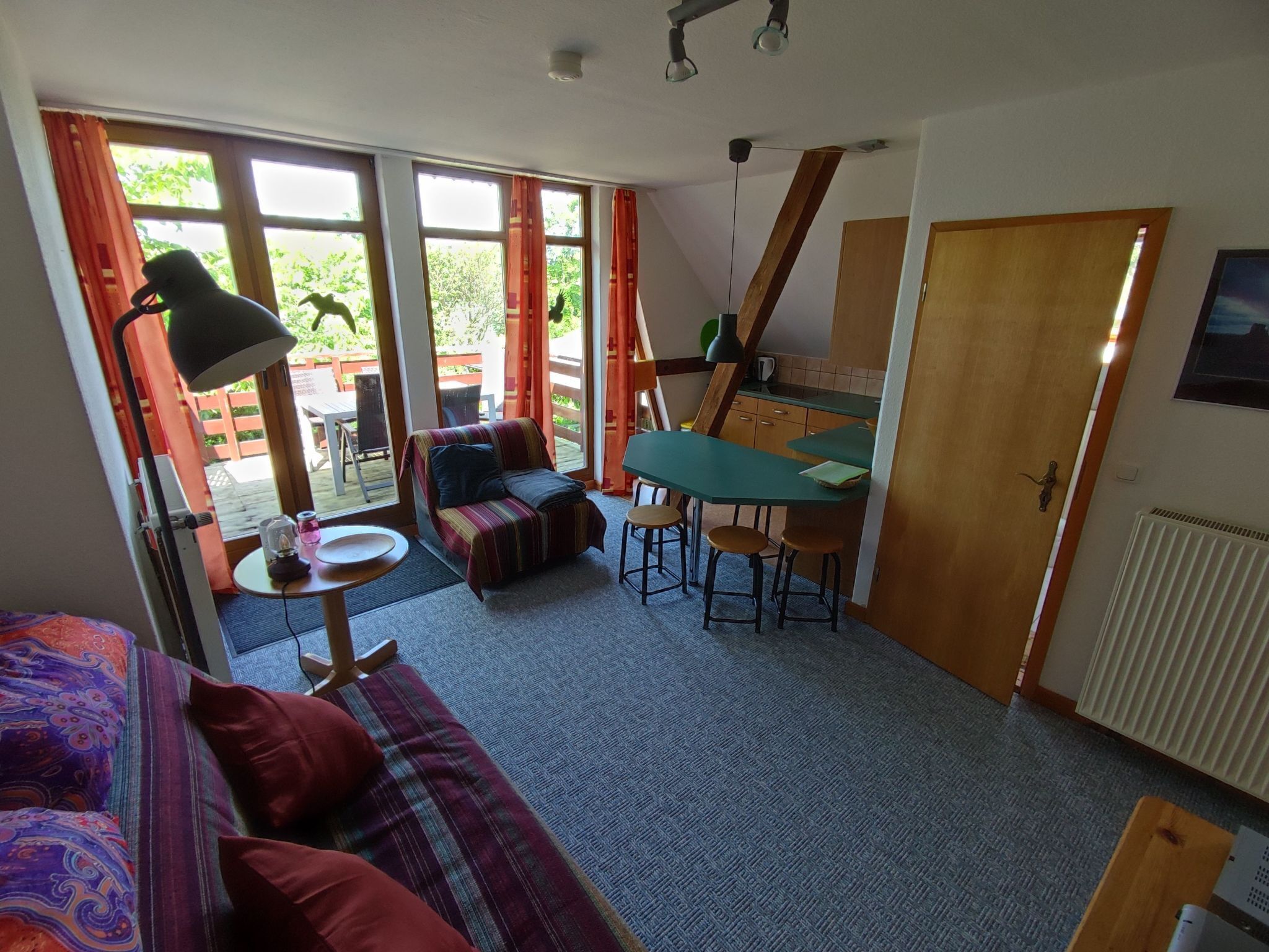 A cozy living room with a sofa, a small table, and a dining area. There are large windows and a door leading to an outdoor area.