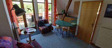 A cozy living room with a sofa, a small table, and a dining area. There are large windows and a door leading to an outdoor area.