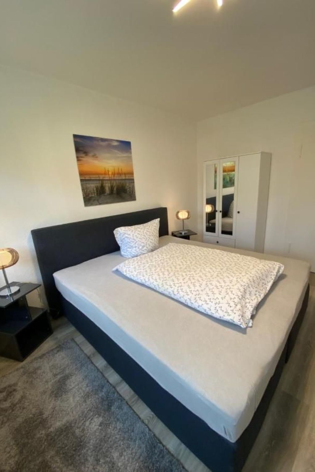 A bedroom with a bed, a nightstand, a lamp, a wardrobe, and a painting on the wall.
