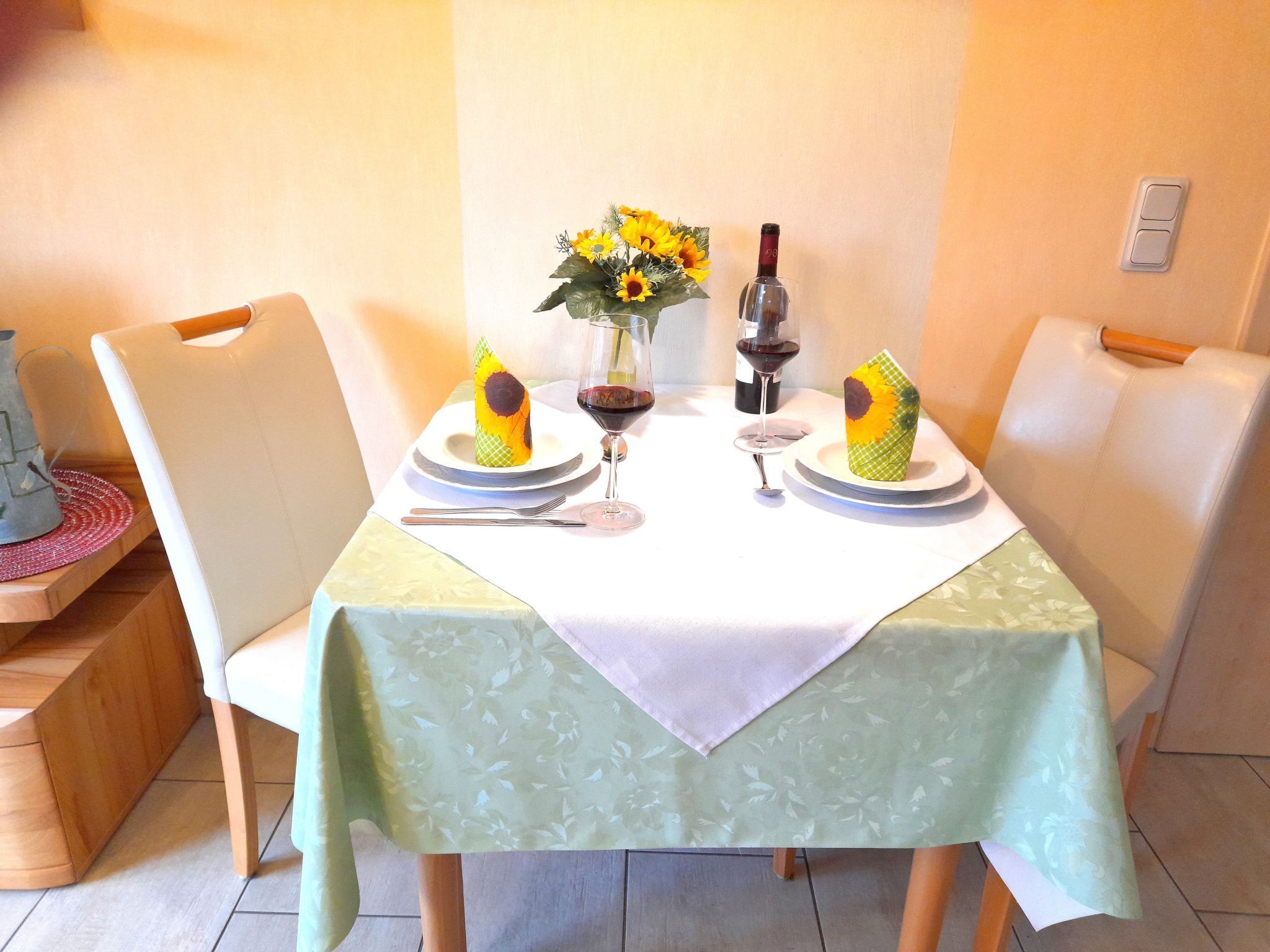 Une table à manger dressée avec des assiettes, des couverts, un vase rempli de tournesols et une bouteille de vin rouge.