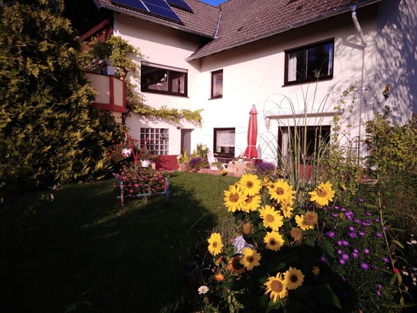 Una casa de dos pisos con un jardín que incluye girasoles y otras flores.