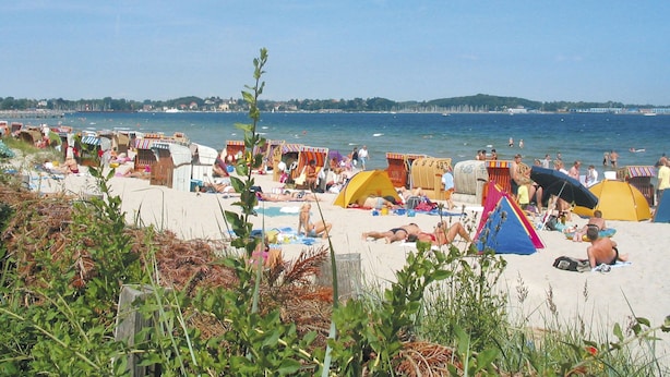 Uma praia com várias tendas e guarda-sóis coloridos, uma faixa de areia e uma vista clara do mar e colinas ao longe.