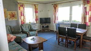 Een woonkamer met een bank, fauteuil, eettafel met stoelen, een televisie en ramen met gordijnen.