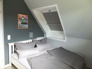 Ein Schlafzimmer mit schrägem Dach, einem Bett mit grauer Bettwäsche, einem Nachttisch, einer Lampe und einem Gemälde von einem Leuchtturm.