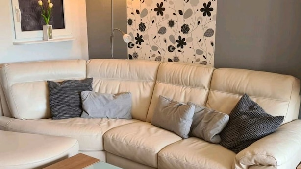 En beige lädersoffa med flera kuddar, ett soffbord i glas och en vägg med ett blommönster.