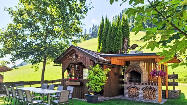 Ein rustikaler Sitzbereich im Freien mit einem Holztisch und -stühlen, einer kleinen Holzhütte mit Schornstein und einer Pergola mit hängenden Blumen.