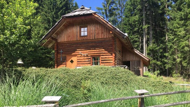 Uma cabana rústica de toras com telhado inclinado, rodeada por uma floresta densa.