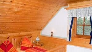 Um quarto de cabana de madeira aconchegante, com teto inclinado, uma cama com roupa de cama colorida, uma mesinha com abajur e uma janela com cortinas.