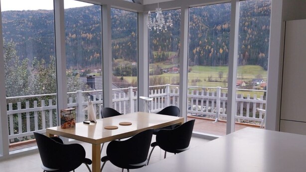 Un comedor con mesa y sillas, ventanas amplias con vistas a un paisaje boscoso y una barandilla blanca en el exterior.